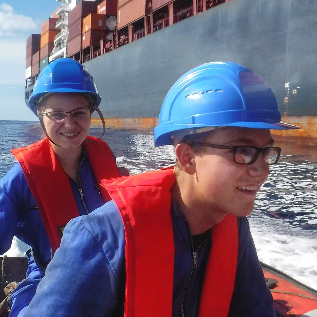 Zwei junge Teilnehmer des VDR-Ferienfahrerprogramms sitzen auf einem kleinen Boot und tragen blaue Schutzhelme sowie rote Schwimmwesten über blauer Arbeitskleidung. Sie lächeln in die Kamera, während im Hintergrund ein großes Containerschiff auf dem Meer zu sehen ist. Das Boot erzeugt weißen Schaum, während es durch das blaue Wasser fährt.