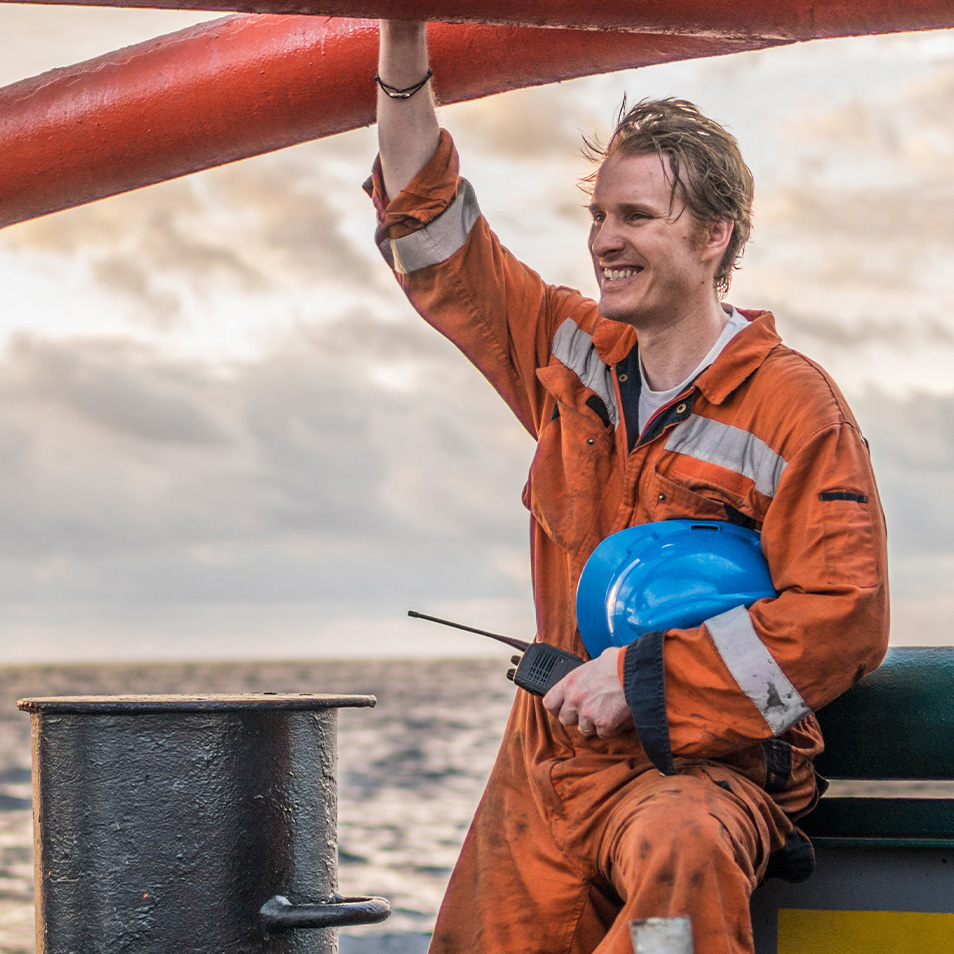 Ein lächelnder Crew-Mitarbeiter in orangefarbener Arbeitskleidung mit Reflektorstreifen hält sich an einem roten Schiffsgeländer fest und trägt einen blauen Schutzhelm und ein Funkgerät, im Hintergrund ist das Meer bei bewölktem Himmel zu sehen