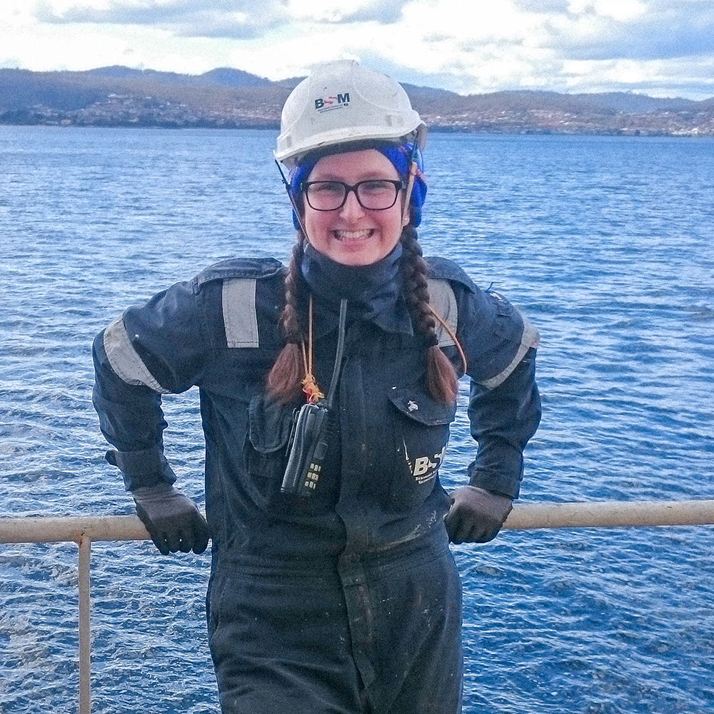 Dana Leathley steht in einem blauen Overall und mit weißem Schutzhelm lächelnd an der Reling eines Schiffs